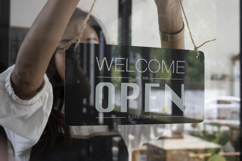 An image of a "We Are Open" sign being hung on a glass door.